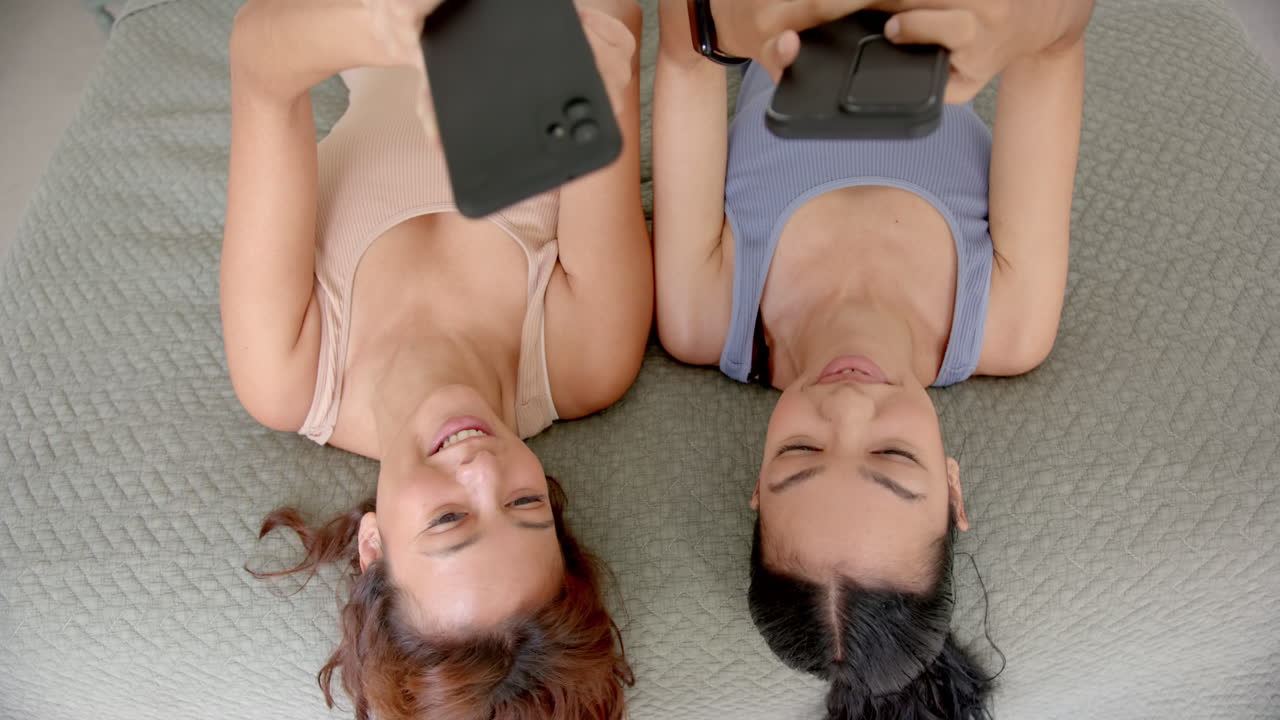 dos jóvenes amigas biraciales se acuestan espaldas a espaldas, sonriendo mientras sostienen teléfonos inteligentes