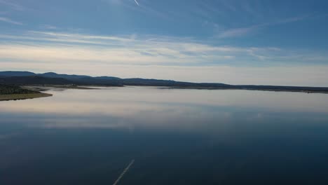 flying-with-a-drone-in-a-reservoir-we-see-the-reflection-of-the-blue-and-white-sky-with-clouds-in-the-water-as-if-it-were-a-mirror-there-is-the-wake-of-a-plane-and-we-see-a-shore-everything-is-calm