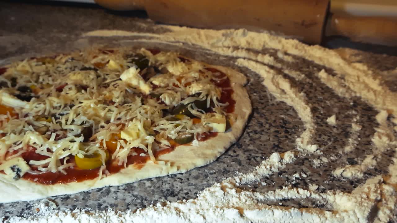 Freshly made raw pizza dough on a home kitchen table with pizza toppings like cheese, meat and tomato sauce on it. White flour around it on the table and a rolling pin in the bokeh blurry background