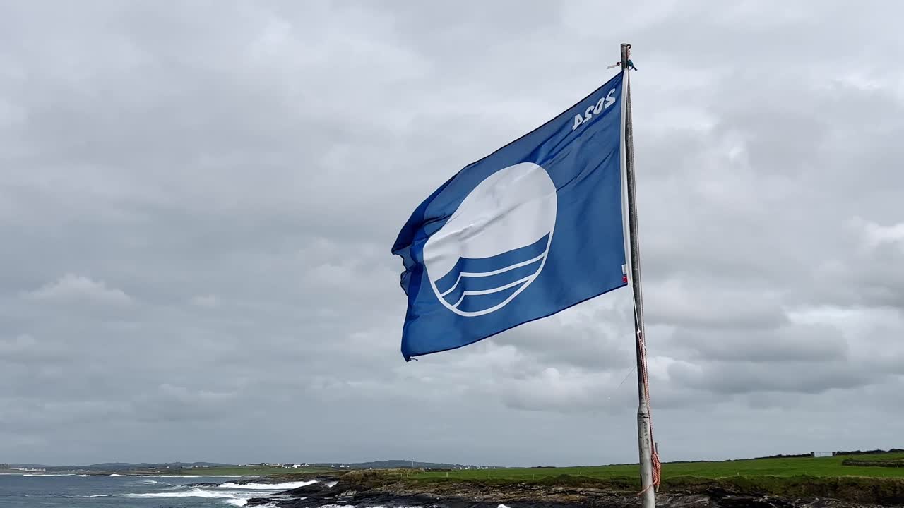 A Blue Flag with ASOS logo waves by a rocky coastline under a cloudy sky