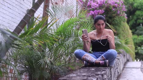 young girl sitting on a stone wall, listening to music from her phone, through a wired headset