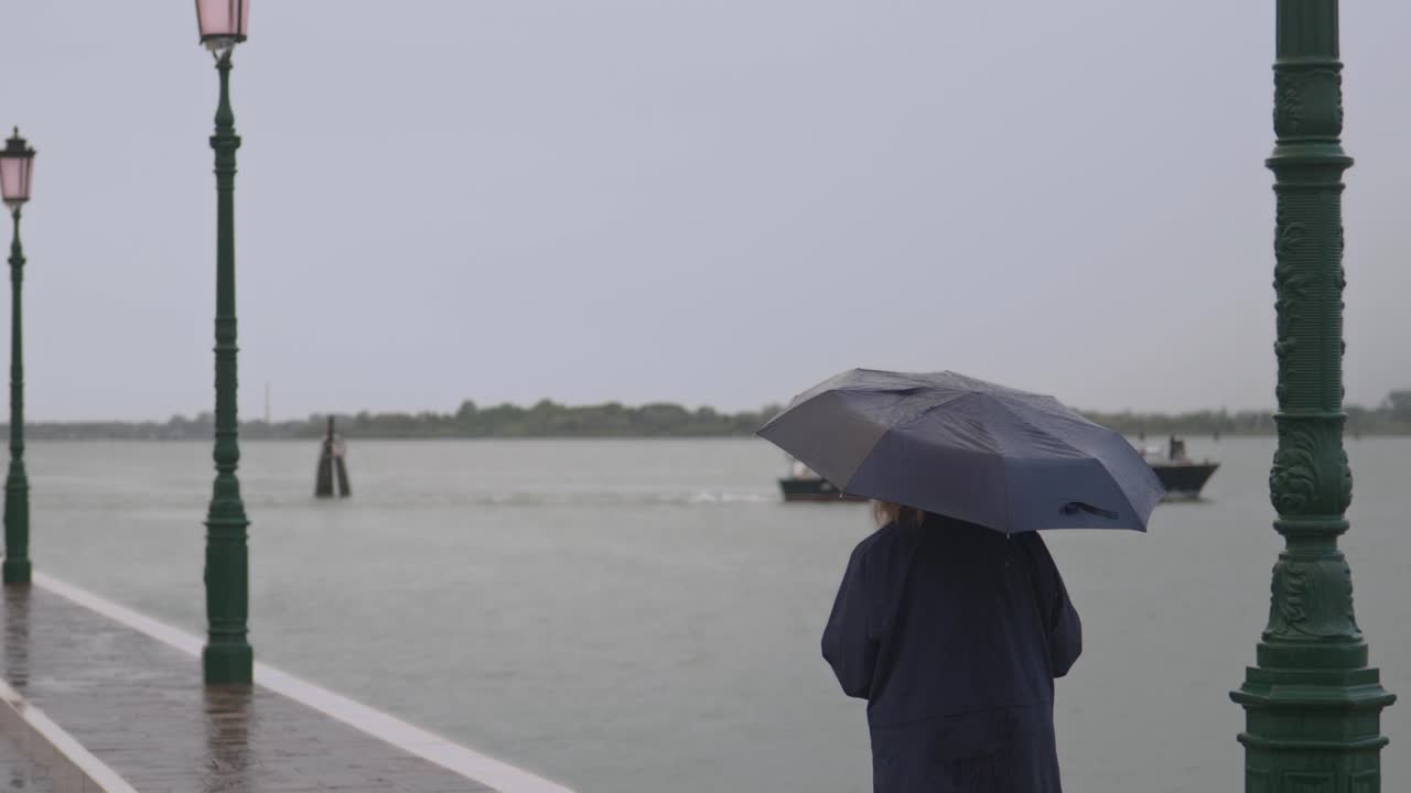 Woman with umbrella stands on waterfront in rain watching passing boat