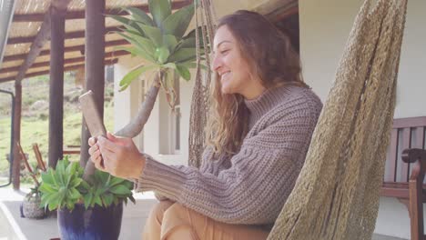 mujer caucásica sonriente acostada en una hamaca usando una tableta en una terraza de jardín rural