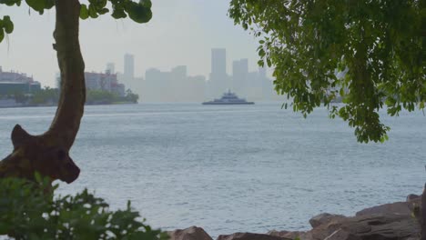 A sleek yacht moves calmly across the water, framed by trees with Miami’s hazy skyline in the backdrop.