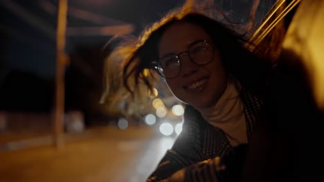una feliz chica morena con gafas redondas y un abrigo blanco y negro mira por la ventana de un coche moderno durante su viaje por la ciudad nocturna. feliz mujer de negocios morena mira feliz desde la ventana del coche por la noche