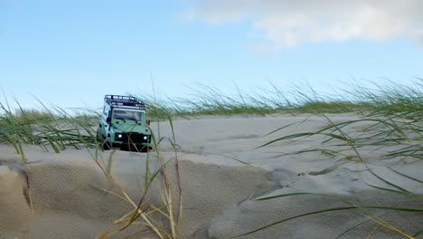 Small-RC-jeep-driving-on-sandy-beach-dunes-against-blue-sky,-static-view