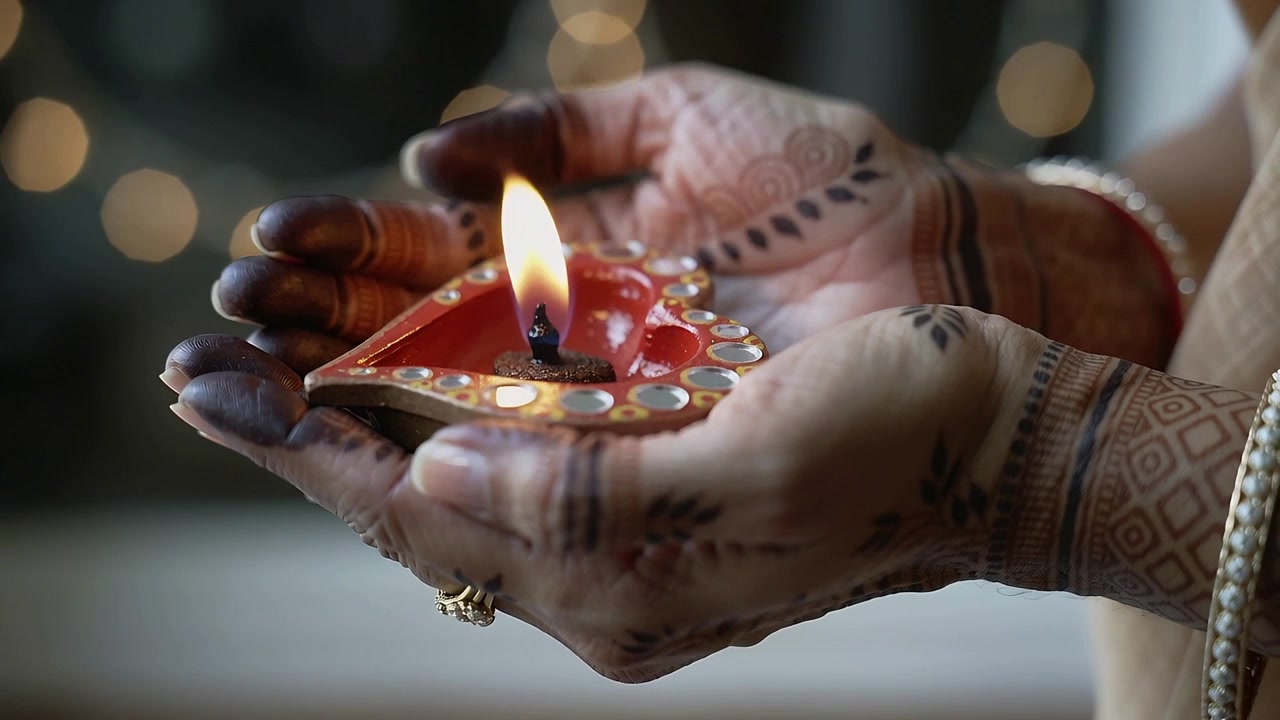 Woman's mehndi hands holding red diya at home, wick lighting, shielding flame with printed sleeve