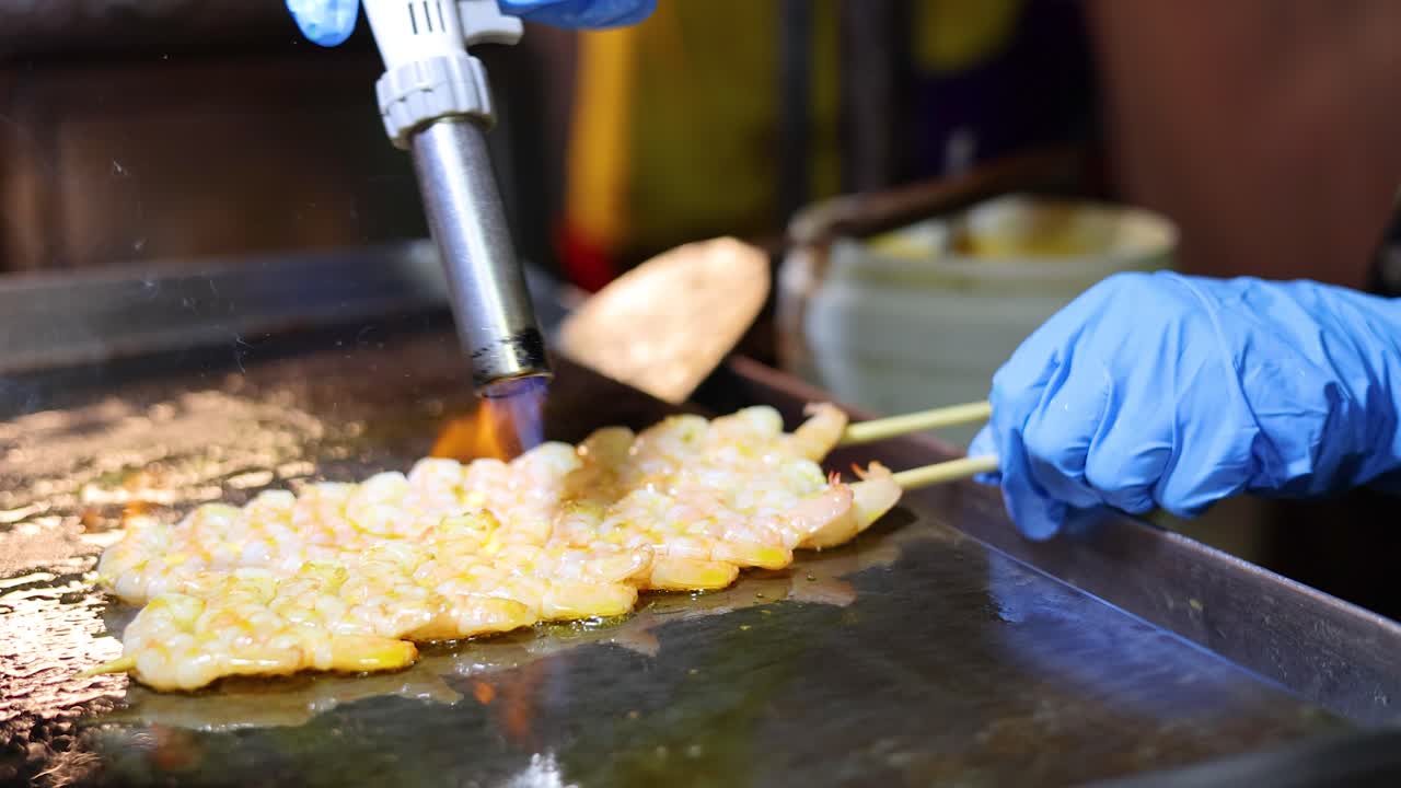 Gloved vendor blowtorches shrimp skewers on flat top at vibrant Bangkok street food market