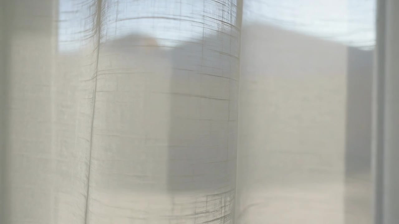 Swaying curtain at window, gentle breeze moving off-white fabric and revealing hill silhouette