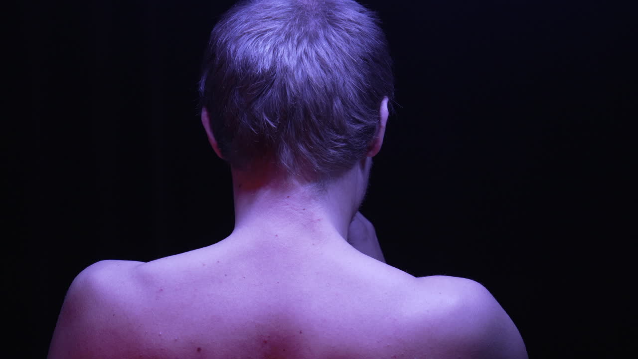 등을 돌린 벌거벗은 남자가 절망에 빠져 머리를 털고 조명 아래서 담배를 피운다