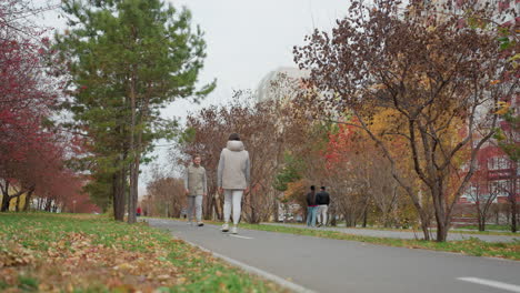 young adults dressed in identical casual outfits walk toward each other along tree-lined path surrounded by colorful autumn foliage as other people stroll nearby