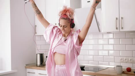 happy young woman with pink hair dancing in kitchen wearing pink pajamas and listening to music with headphones