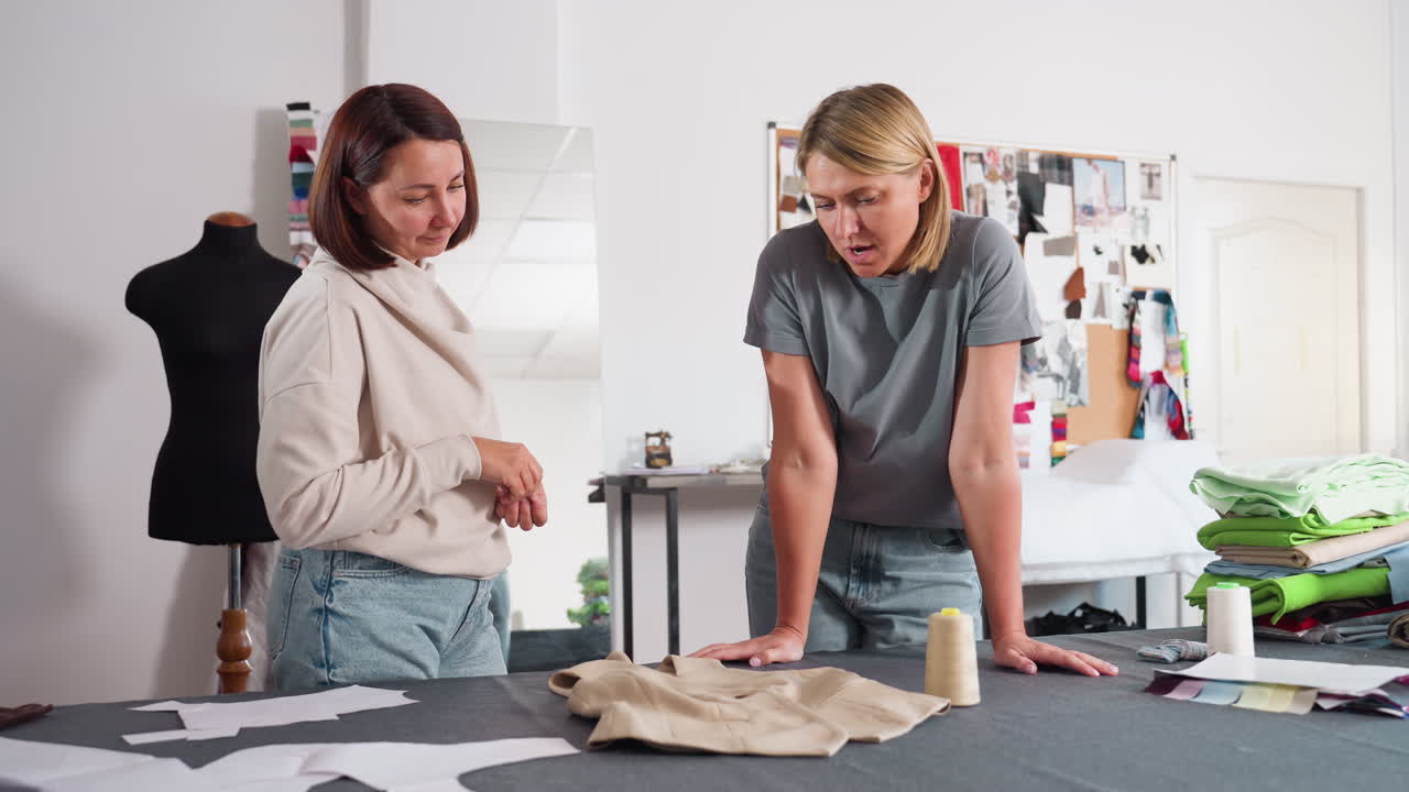Dressmakers collaborating around jacket, placing buttons, giving supportive thumbs up, showcasing teamwork, craftsmanship, pattern pieces, spools, mannequin and inspiration board in fashion workspace