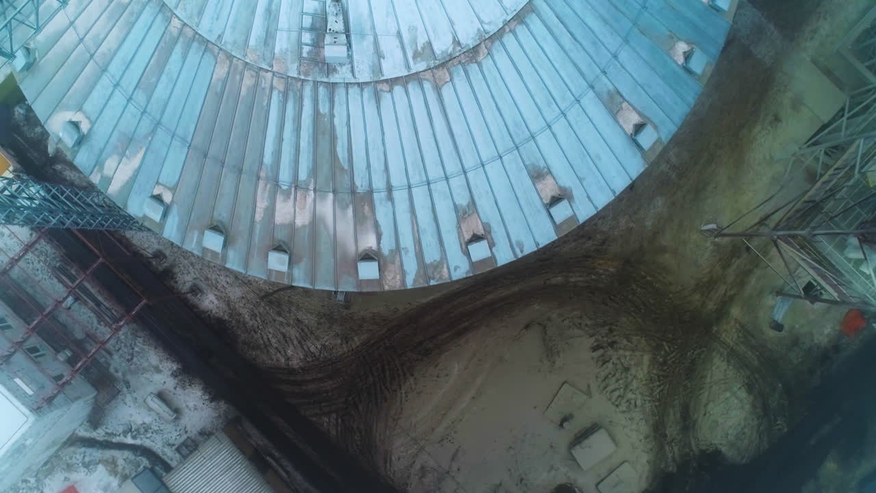 Round metal roof of silo tank. Flying over the grain container. Dirty land covered with dirty snow and trails of car wheels. Top view.