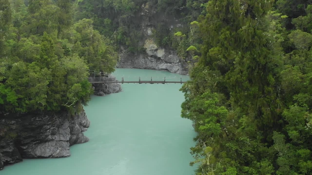 SLOWMO - Rekaman drone udara jembatan gantung di atas sungai gletser biru di Ngarai Hokitika, Selandia Baru