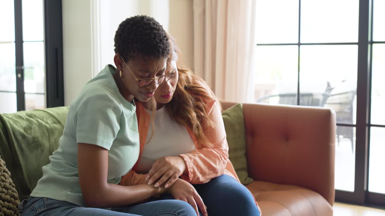 Female friends upset holding head comforter in ripped jeans leaning, hugging on tan sofa at home