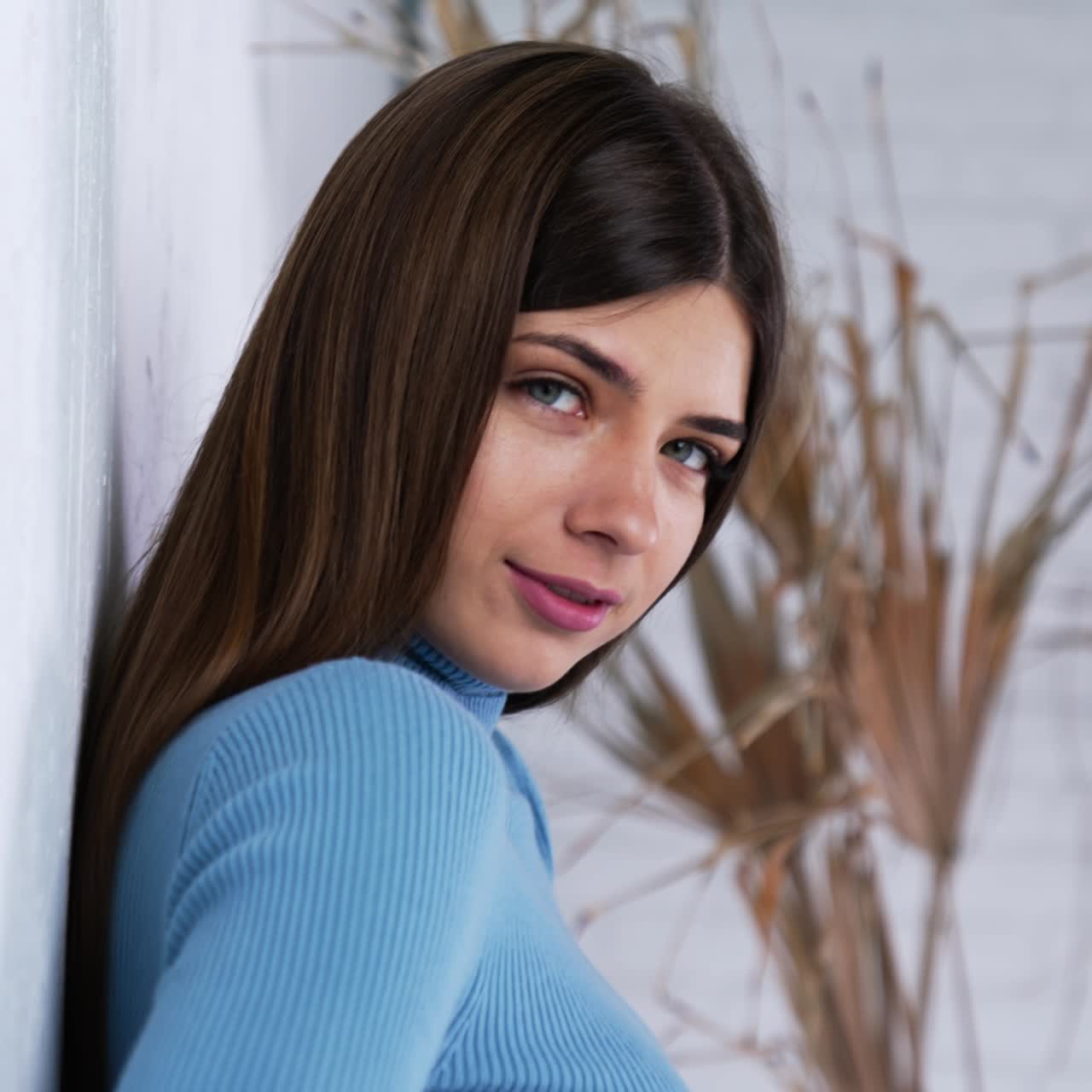 Young dark-haired lady with no make-up wearing blue clothes. Attractive girl standing at the wall smiling to the camera. Close up