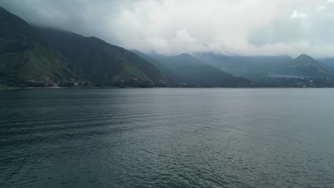 Drone-view-in-Guatemala-flying-over-a-blue-lake-surrounded-by-green-mountains-and-volcanos-on-a-cloudy-day-in-Atitlan