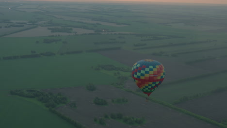 colorful checkered hot air balloon carrying passengers drifts above endless farmland at dusk, aerial perspective showcasing patchwork fields and distant river under pastel sky