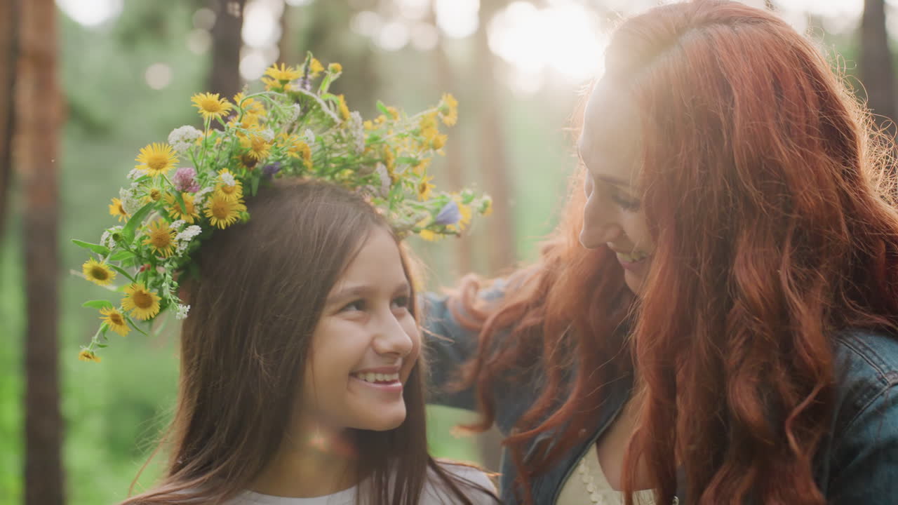 Joyful mom decorates daughter head with bright flowers while girl covers eyes with hands, smiling playfully under soft sunlight in green forest, showing warmth, and tender family bonding outdoors