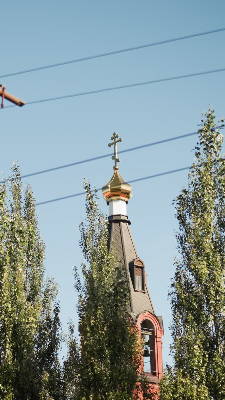 황금 십자가가 있는 교회 첨탑, 맑고 푸른 하늘 아래 붉은 벽돌로 된 종탑을 둘러싼 미루나무, 보이는 전선, 따뜻한 여름 햇살이 화려한 돔과 외관 디테일을 강조하고, 고요한 도시 풍경