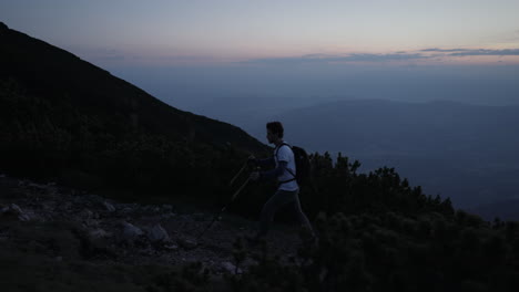 Kamera-Tracking-Wanderer-Von-Der-Seite,-Der-Mit-Wanderstöcken-In-Der-Morgendämmerung-Auf-Einem-Felsigen-Weg,-Der-Von-Nadelbäumen-Umgeben-Ist,-Einen-Hügel-Hinaufgeht