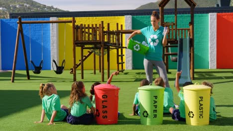 vista frontal de una maestra caucásica que enseña a los escolares sobre el reciclaje de botellas en el patio de recreo escolar