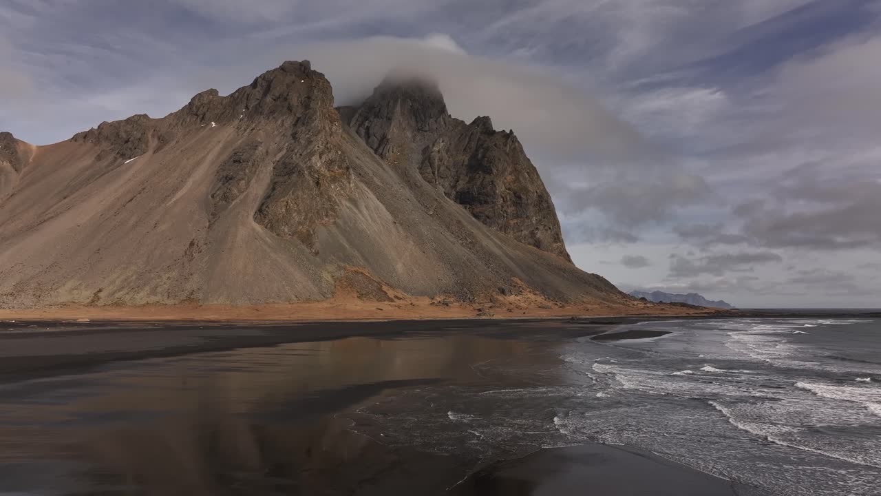 Vestrahorn’s steep ridges loom over black sands and foamy shoreline in dramatic coastal view. Stokksnes, Iceland