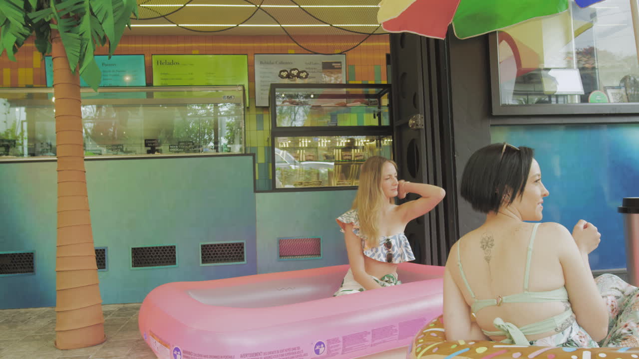 Two young women enjoy ice cream in a relaxed atmosphere next to an inflatable pool in front of a contemporary ice cream parlor.
