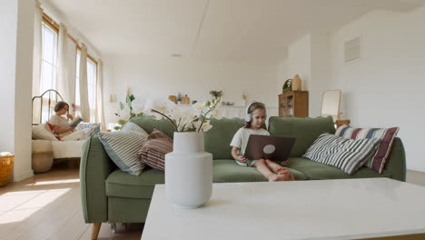 weitwinkelaufnahme eines verantwortungsvollen kleinen blonden mädchens, das ihren online-unterricht in ihrem wohnzimmer erhält, während ihre mutter dahinter ein buch liest