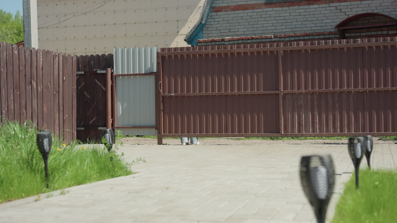 Two women walk out of compound through open gate onto paved walkway in sunny suburban yard, carrying shopping bag and chatting as they pass wooden swing under clear blue sky
