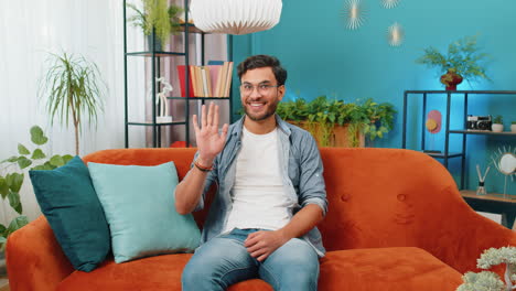 indian man smiling friendly at camera waving hands gesturing hello hi greeting at home on sofa