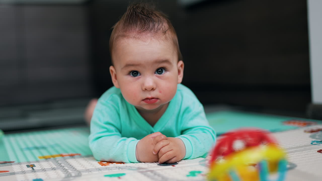 Amazing adorable kid with funny hair lies on belly leaning on elbows. Cute kid stars to laugh banging hands and feet over floor.