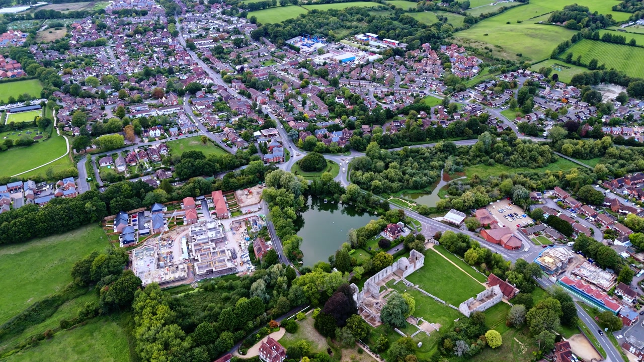 High-angle aerial drone pans left over Bishop’s Waltham village town in England, showcasing houses, winding roads, and autumn trees glowing in warm golden sunset light across the countryside landscape