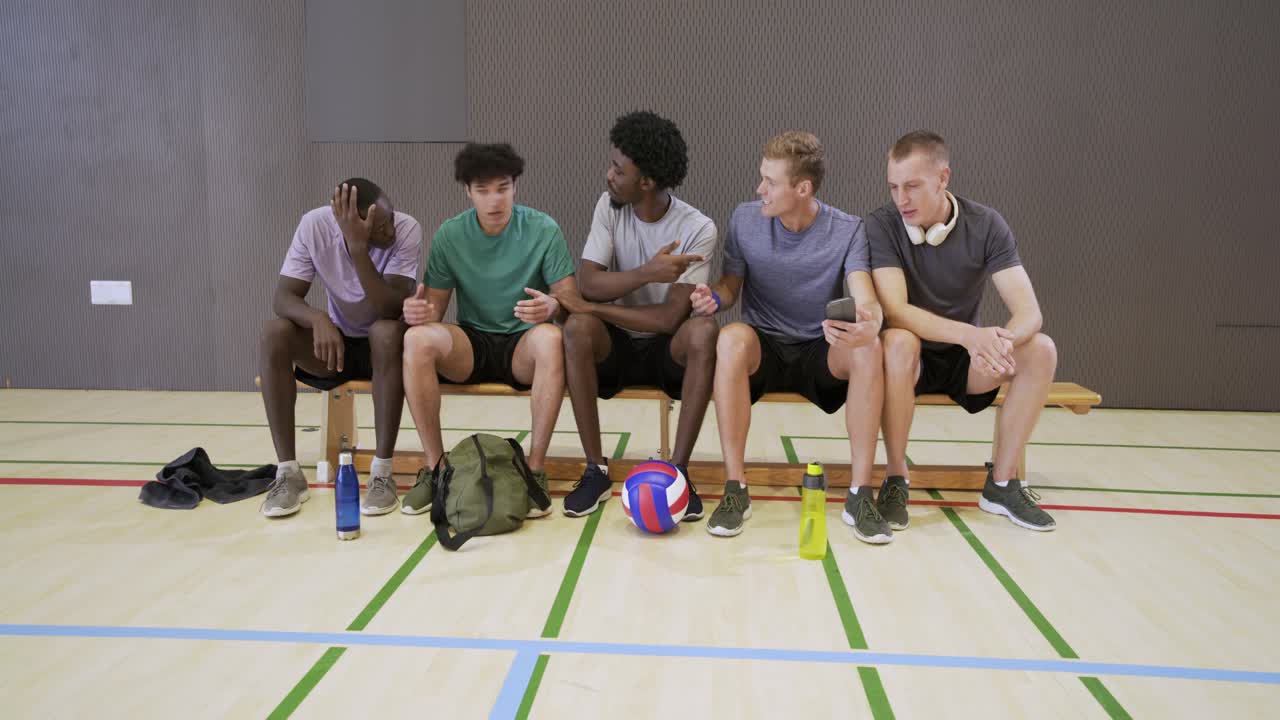 Fünf männliche Teamkollegen sitzen auf einer Turnbank, ein Blonder hält ein Telefon und zeigt einen Clip, ein Volleyball liegt auf dem Boden.