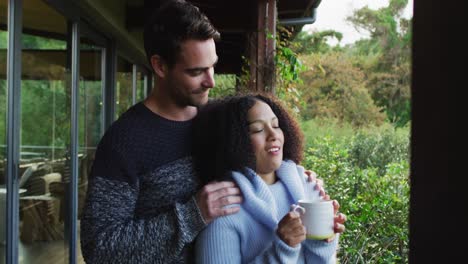 happy diverse couple embracing and drinking coffee standing on balcony in the countryside