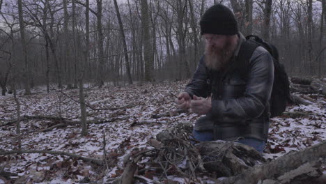 a bearded man in a plaid flannel shirt kneels as he is breaking up sticks to use as kindling for a campfire in a wooded area with a dusting of snow on the ground