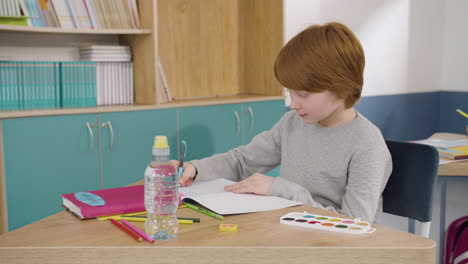 niño de jengibre concentrado sentado en el escritorio y escribiendo en un cuaderno durante la clase de inglés en la escuela