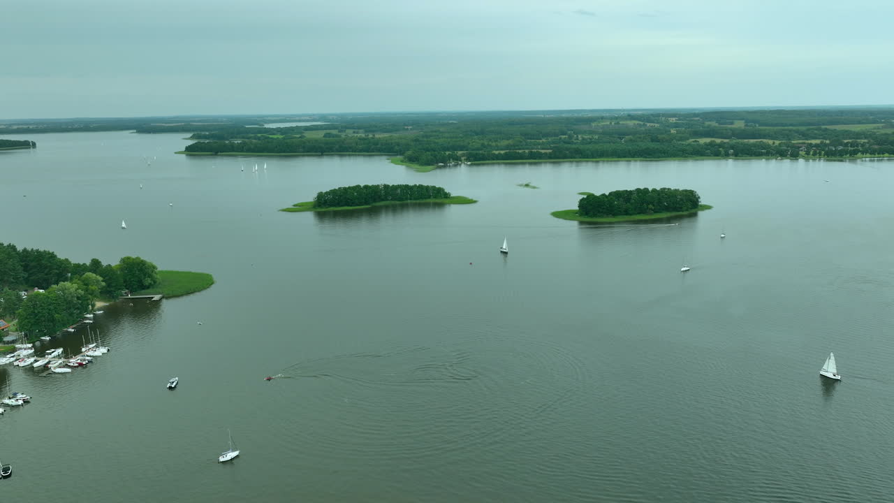 Aerial view of a calm lake with small islands and sailboats, surrounded by green fields and forest - Jeziorak lake, Siemiany