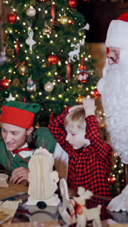 cheerful children are playing with wooden toys with the help of elves and santa claus. two elves in green costumes, santa and little children on the background of christmas tree at home. vertical video