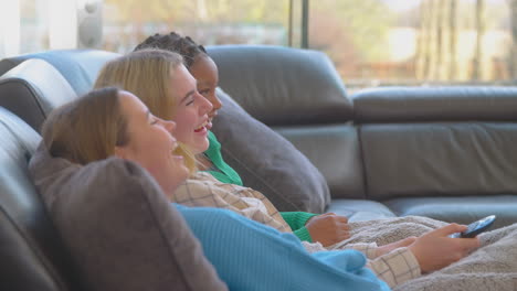 group of multi-cultural teenage girl friends snuggled under blanket watching tv at home