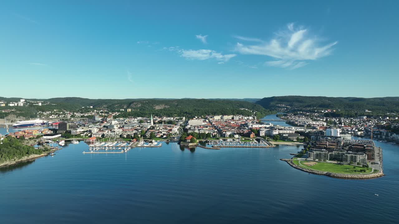 Kristiansand in Norway seen from above, with boats dotting the harbor and green hills framing the waterfront. The blue sky adds to the serene atmosphere on a sunny day