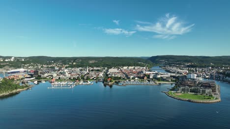 kristiansand in norway seen from above, with boats dotting the harbor and green hills framing the waterfront. the blue sky adds to the serene atmosphere on a sunny day