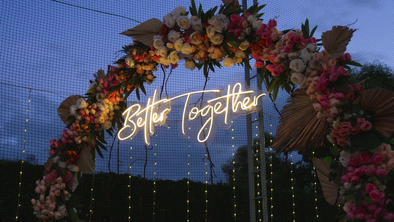 Insegna al neon luminosa "Better Together" appesa a un lussureggiante arco floreale durante una celebrazione pre-matrimoniale indiana sangeet all'aperto al crepuscolo.