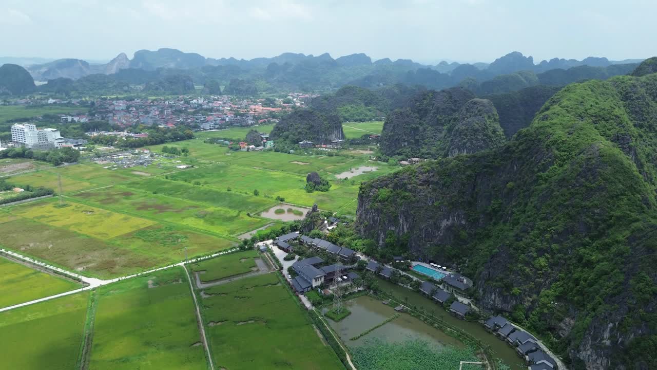Slow ascending drone shot revealing extensive rice fields bordered by limestone formations and distant buildings