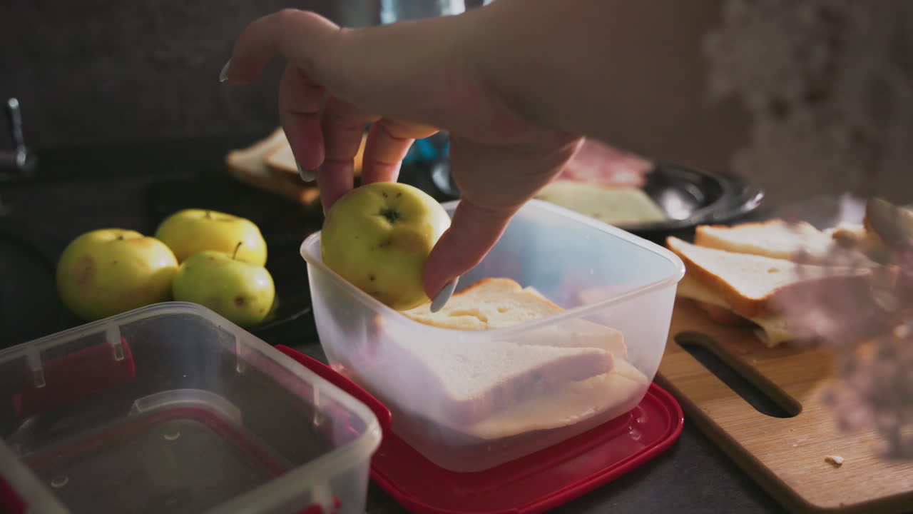Caregiver places sandwich inside plastic container while preparing school lunch on kitchen counter, surrounded by apples, bread slices, and other food, capturing hands in action during early morning routine