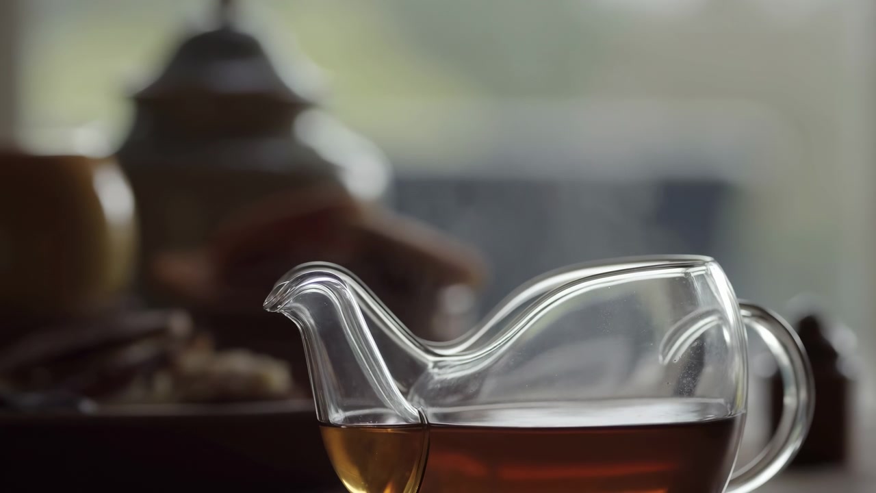 Una tazza di vetro trasparente di tè ambrato si trova in primo piano accanto a una teiera e dei pasticcini, con una morbida luce naturale che entra da una finestra luminosa in un accogliente ambiente interno