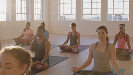 Clase-De-Yoga-De-Jóvenes-Sanos-Practicando-Meditación-En-Postura-De-Loto-Disfrutando-De-Un-Relajante-Ejercicio-De-Respiración-Con-Un-Instructor-En-El-Gimnasio-Al-Amanecer