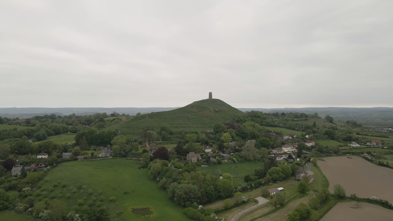 Aerial view of Glastonbury, drone rotating and camera facing down over the Glastonbury Tor, drone flying over the green fields with some houses, some trees on the left side of the camera. 4K, 60fps.