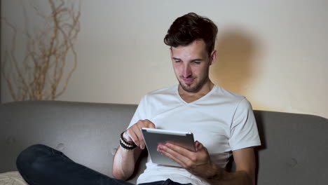 Un-Joven-Con-Barba-Y-Un-Peinado-Moderno,-Vestido-Con-Una-Camiseta-Blanca-Y-Jeans,-Sentado-En-Un-Sofá-En-Casa,-Con-Las-Piernas-Cruzadas,-Asintiendo-Con-La-Cabeza-En-Señal-De-Aprobación-Mientras-Pasa-El-Dedo-Por-Su-Tableta,-Estático-4k